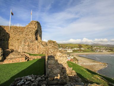 A stone castle ruin on a grassy hill with flags flying and a coastal town with a beach and hills in the background at Llwyn Onn in Criccieth