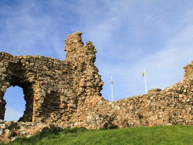 Ruins of a stone castle with two flagpoles displaying flags on a grassy hill at Llwyn Onn in Criccieth
