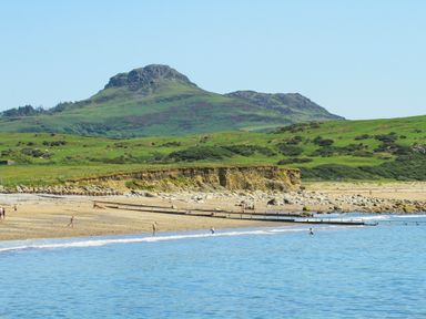 A beach with several people near the water and a green hill with rocky peaks in the background at Llwyn Onn in Criccieth