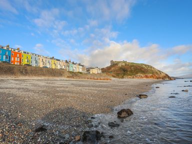 A pebble beach with rocks near the water and a row of colorful houses along a stone wall with a hill and castle ruins in the background at Llwyn Onn in Criccieth