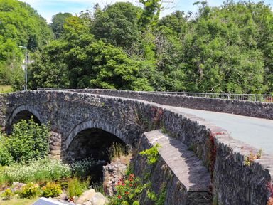 A stone arch bridge over a small waterway surrounded by green trees and vegetation at Llwyn Onn in Criccieth