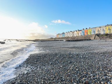 A pebble beach with ocean waves and a row of colorful houses on a seawall at Llwyn Onn in Criccieth