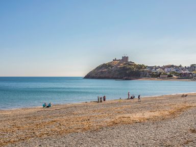 A beach with people sitting and walking near water and a hill with a castle and houses in the background at Llwyn Onn in Criccieth