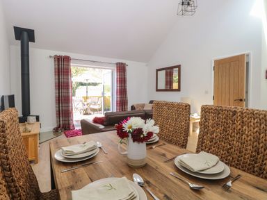 A dining area with a wooden table set with plates and napkins and a living room with a brown sofa and glass doors opening to an outdoor seating area at Beck View in Weston Longville near Lenwade
