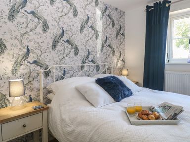 A bedroom with a bed with white bedding a tray with pastries orange juice and a newspaper a peacock wallpaper and blue curtains at Beck View Weston Longville near Lenwade