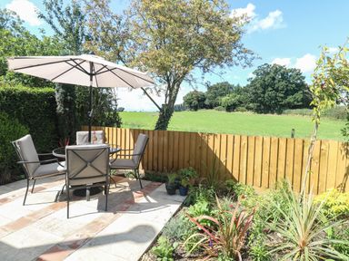 A garden patio with a table chairs and umbrella next to a wooden fence and green field at Beck View in Weston Longville near Lenwade