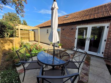 An outdoor patio area with a round glass table and four chairs next to a brick building with double glass doors at Beck View in Weston Longville near Lenwade