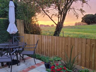 A patio area with a glass table and four chairs next to a wooden fence and plants at Beck View in Weston Longville near Lenwade