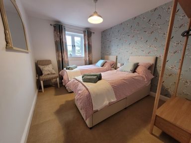 A bedroom with two single beds towels and pillows near a window with curtains a striped chair and floral wallpaper at Beck View Weston Longville near Lenwade