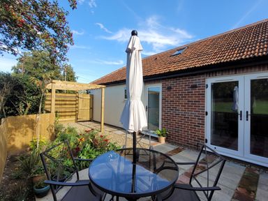 An outdoor patio with a glass table and four chairs with a closed umbrella in a garden at Beck View in Weston Longville near Lenwade
