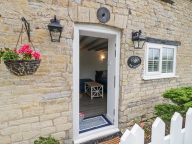An open door with a view of a living room through a stone wall exterior with hanging flower basket and two black lanterns at The Cottage in Ettington
