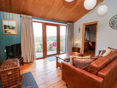 A living room with a brown sofa a wooden coffee table a television and glass doors opening to a balcony at Y Caban Glas in Rhydlewis near Brynhoffnant