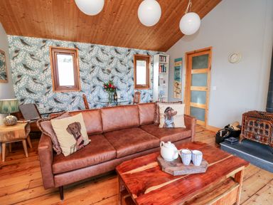 A living room with a brown sofa with dog print cushions a wooden coffee table with a teapot and cups and a wood stove at Y Caban Glas in Rhydlewis near Brynhoffnant