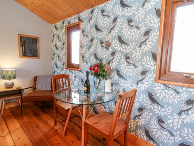 A dining area with a glass table two wooden chairs a bouquet of flowers a bottle and two wine glasses next to a chair with a lamp on a side table and patterned bird wallpaper at Y Caban Glas Rhydlewis near Brynhoffnant