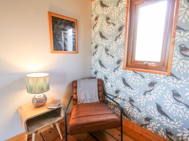 A corner with a brown chair with a gray throw a wooden side table with a lamp and a book and a window with bird patterned wallpaper at Y Caban Glas in Rhydlewis near Brynhoffnant