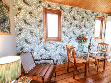 A dining area with a glass table and wooden chairs and a brown armchair against a wall with bird wallpaper at Y Caban Glas in Rhydlewis near Brynhoffnant