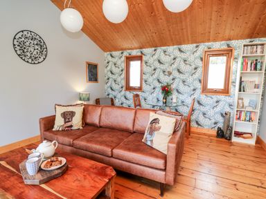 A living room with a brown leather sofa with animal print cushions a wooden coffee table with a tea set a glass dining table with chairs and a bookshelf at Y Caban Glas Rhydlewis near Brynhoffnant