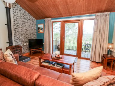A living room with a brown sofa wooden coffee table television wood stove and glass doors leading to a balcony at Y Caban Glas in Rhydlewis near Brynhoffnant