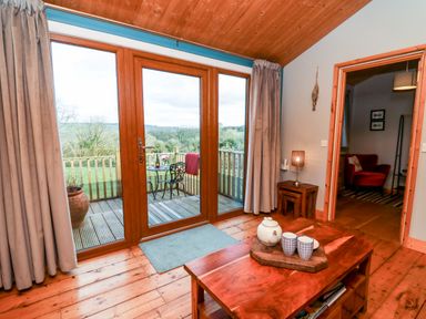 A living room with a wooden coffee table and glass doors opening to a balcony with outdoor furniture at Y Caban Glas in Rhydlewis near Brynhoffnant