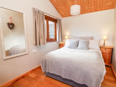 A bedroom with a bed covered in grey blankets and pillows two wooden bedside tables with lamps a window with curtains and a wall mirror at Y Caban Glas in Rhydlewis near Brynhoffnant