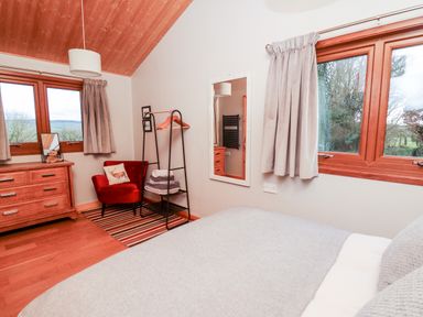 A bedroom with a bed a red armchair a wooden dresser and windows with curtains at Y Caban Glas in Rhydlewis near Brynhoffnant