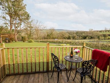 A small deck with a metal table and two chairs with wine glasses and a bottle overlooking a grassy yard and trees at Y Caban Glas in Rhydlewis near Brynhoffnant