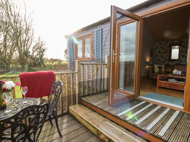 A small outdoor wooden deck with a metal table and chairs next to an open wooden door leading to a living room at Y Caban Glas in Rhydlewis near Brynhoffnant