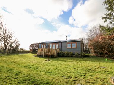 A small grey house with wooden framed windows and a wooden deck on a grassy lawn surrounded by trees at Y Caban Glas in Rhydlewis near Brynhoffnant