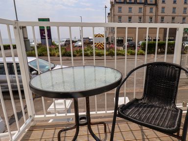 A small balcony with a glass table and a black woven chair overlooking a street with cars and a large building at Beach Bridlington
