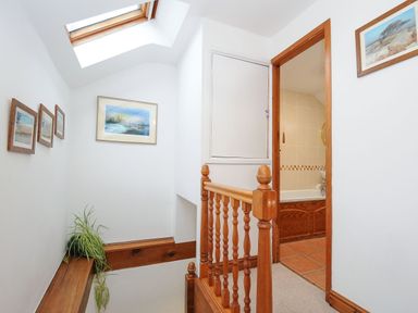 A hallway with wooden banisters and framed pictures on white walls with a skylight window and a doorway leading to a tiled bathroom at Henwood Barn in Henwood near Upton Cross