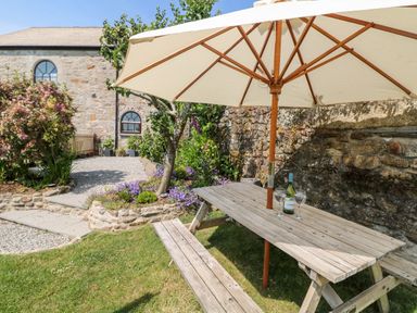 An outdoor garden area with a wooden picnic table and umbrella a bottle and two glasses on the table and stone building in background at Henwood Barn in Henwood near Upton Cross