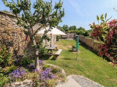 A garden with a tree and flowers around it a picnic table with an umbrella a stone wall and grass at Henwood Barn Henwood near Upton Cross
