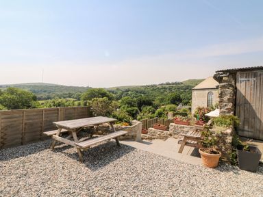 An outdoor patio area with wooden picnic table and benches plants in pots and a stone wall with countryside view at Henwood Barn in Henwood near Upton Cross