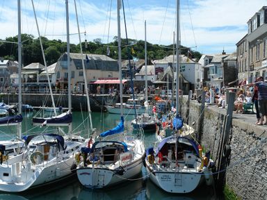 A harbor with several sailboats docked near a stone wall and people gathered along the waterfront in front of buildings at Mole Cottage in Henwood near Upton Cross