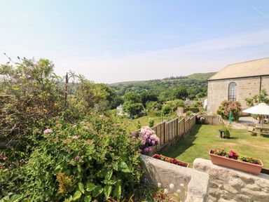A garden with flowers and grass next to a stone building and wooden fence at Mole Cottage in Henwood near Upton Cross