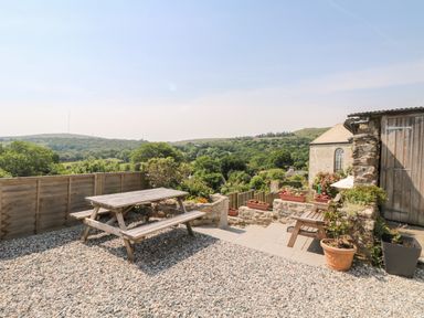 An outdoor seating area with a wooden picnic table and benches and a stone patio with potted plants overlooking green hills at Mole Cottage in Henwood near Upton Cross