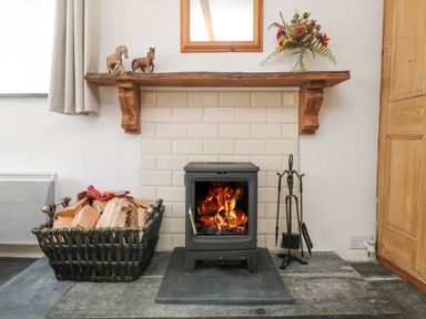 A wood burning stove with fire inside a tiled fireplace with a basket of firewood on the left and fireplace tools on the right at Mole Cottage in Henwood near Upton Cross