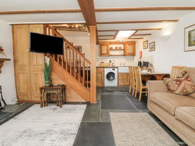 A living room with a beige sofa pillows and wooden dining table next to a kitchen with wooden cabinets washing machine and stairs with a mounted TV at Mole Cottage Henwood near Upton Cross