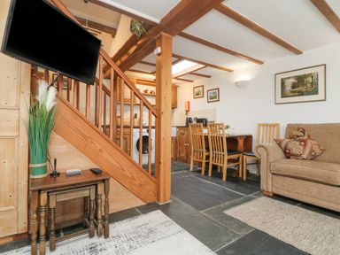 A living area with wooden stairs a mounted television a small table four dining chairs a kitchen with appliances and a sofa with cushions at Mole Cottage in Henwood near Upton Cross