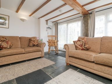 A living room with two beige sofas and patterned cushions a wooden rocking chair and large windows at Mole Cottage in Henwood near Upton Cross