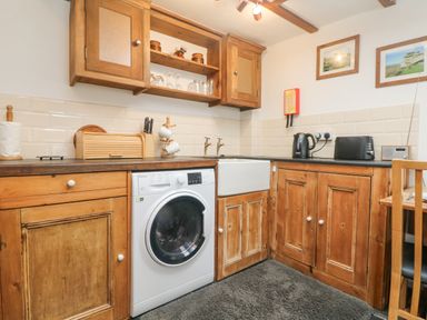 A kitchen with wooden cabinets a washing machine a white sink a kettle a toaster and framed pictures on the wall at Mole Cottage Henwood near Upton Cross