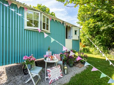 A small outdoor seating area with a table, chair, flowers, and bunting outside a blue shepherds hut in a garden at Gwithian Shepherds Hut in Angarrack near Hayle