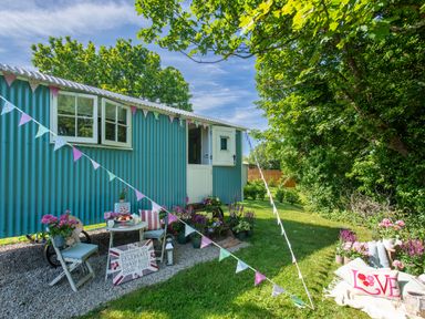 An outdoor garden area with a teal shepherds hut a small table and chairs with flowers at Gwithian Shepherds Hut Angarrack near Hayle