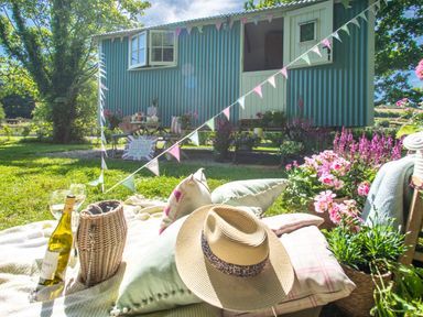 A picnic setup with cushions a straw hat a bottle of wine and glasses on grass near a blue shepherds hut with flowers at Gwithian Shepherds Hut Angarrack near Hayle