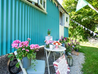 A small outdoor seating area with a table, potted flowers, a lantern, and a teal-colored building in the background at Gwithian Shepherds Hut in Angarrack near Hayle