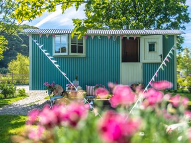 A small blue corrugated metal hut with bunting and outdoor chairs and table set among trees and flowers at Gwithian Shepherds Hut in Angarrack near Hayle