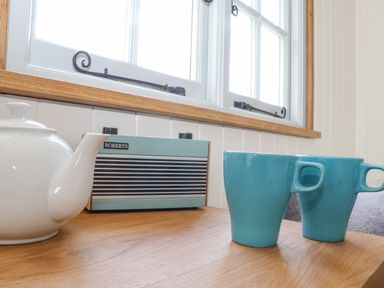 A white teapot two blue mugs and a vintage radio on a wooden table near a window at Gwithian Shepherds Hut in Angarrack near Hayle