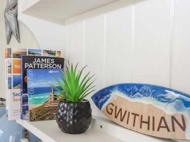 A shelf with books, a small plant in a textured black pot, and a wooden sign with blue resin and the word GWITHIAN at Gwithian Shepherds Hut in Angarrack near Hayle