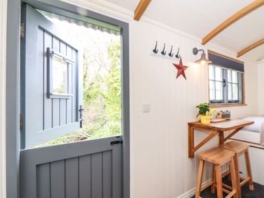 A small room with a blue stable door a wooden table and stools a window and coat hooks at Godrevy Shepherds Hut Angarrack near Hayle