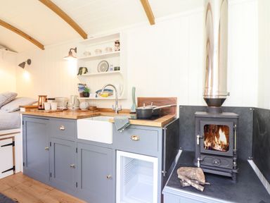A kitchen area with blue cabinets a sink a stove and a wood burning stove with fire at Godrevy Shepherds Hut Angarrack near Hayle